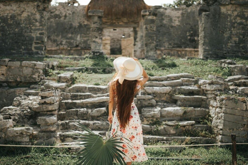 A stylish woman in a floral ANTHROPOLOGIE dress and wide-brimmed hat stands with her back to the camera, gazing at ancient stone ruins. She holds a large green leaf and touches her hat, enveloped by lush greenery and a sunlit sky.