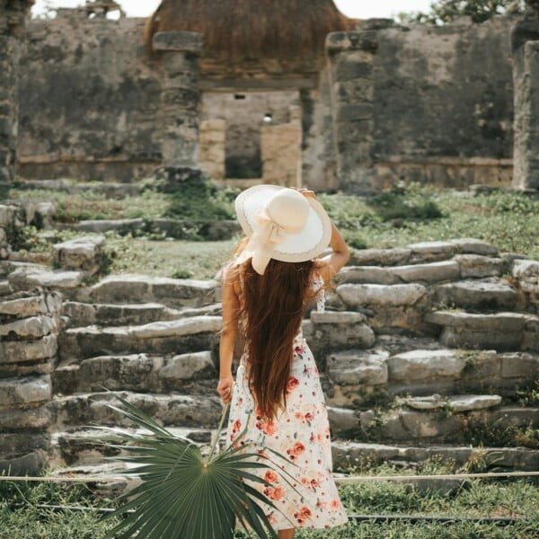 5 Stylish Modest ANTHROPOLOGIE Dresses - Under $200 11 A stylish woman in a floral ANTHROPOLOGIE dress and wide-brimmed hat stands with her back to the camera, gazing at ancient stone ruins. She holds a large green leaf and touches her hat, enveloped by lush greenery and a sunlit sky.