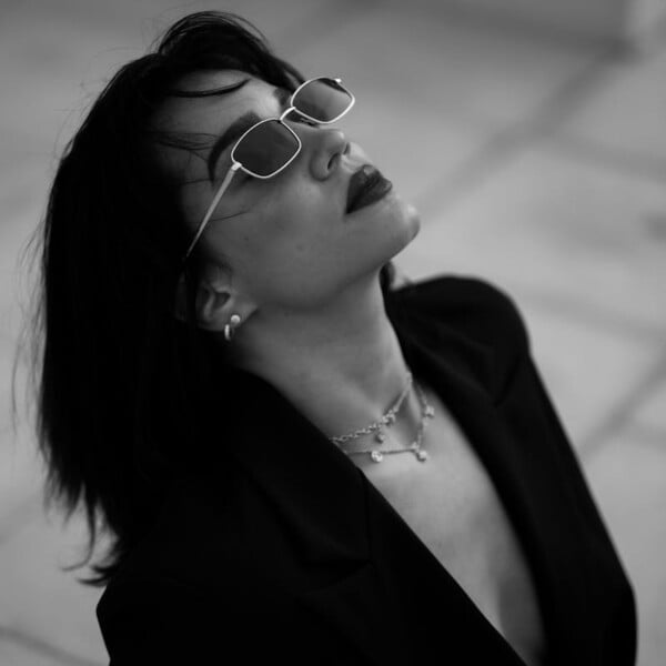 Celebrity inspired looks for over-40-women 12 A black and white photo captures a person channeling celebrity-inspired looks with sunglasses and a blazer, eyes lifted. Their shoulder-length hair complements a chic necklace against the blurred, tiled background. A nod to fashion trends embraced by over-40 women.