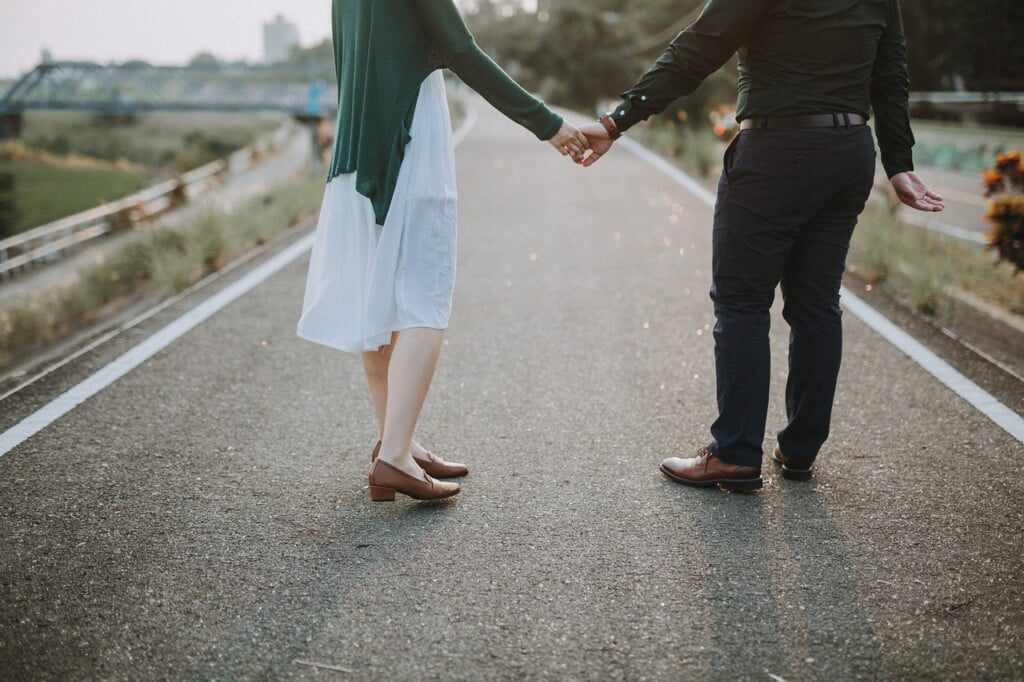 A couple strolls hand in hand down an empty road, the woman confidently flaunting a classic date night outfit—a white dress with a green sweater. The man complements her style in dark pants and a black shirt. Theyre surrounded by lush greenery with a distant bridge adding to their serene journey.