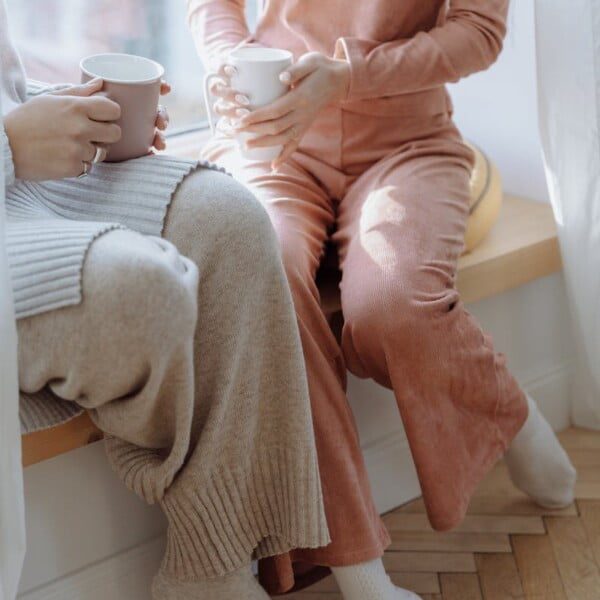 Two people sit on a window ledge in cozy outfits, holding mugs. One wears a chic beige sweater with matching pants, the other in a pink loungewear set. Natural light filters through sheer curtains, creating a warm, relaxed atmosphere perfect for working from home.
