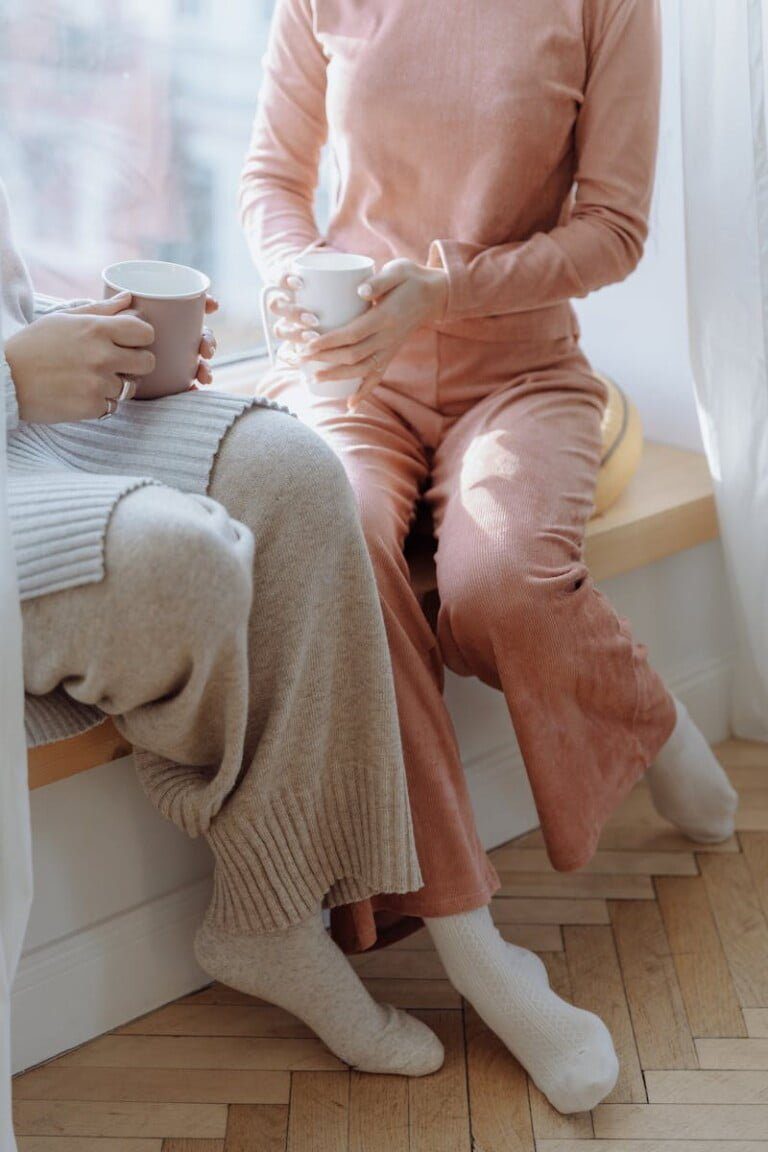 Look Chic Even When Working From Home! 1 Two people sit on a window ledge in cozy outfits, holding mugs. One wears a chic beige sweater with matching pants, the other in a pink loungewear set. Natural light filters through sheer curtains, creating a warm, relaxed atmosphere perfect for working from home.