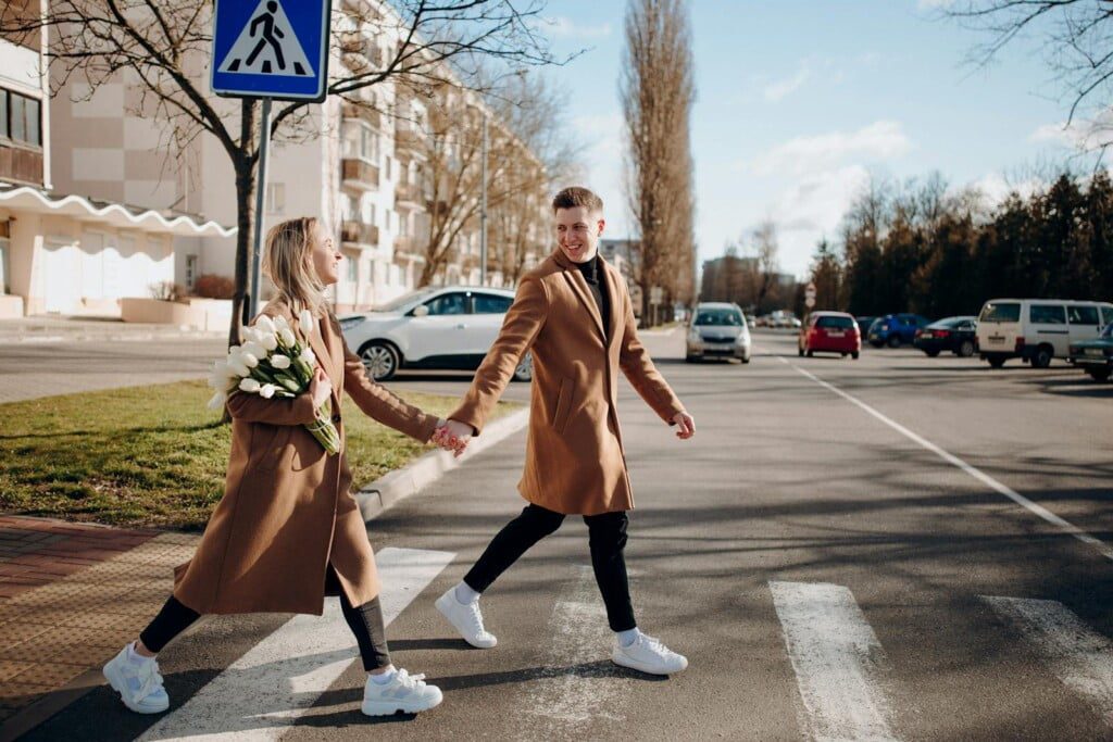 A smiling couple wearing matching stylish trench coats and white sneakers hold hands and cross a street on a sunny day. The woman carries a bouquet of white tulips. There are cars and buildings in the background.