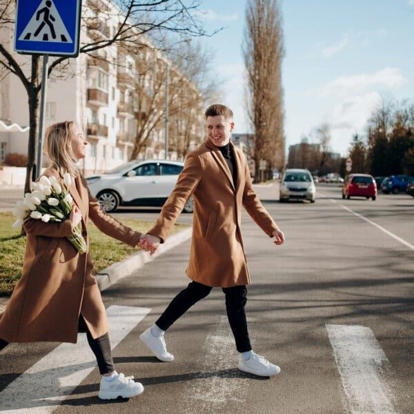A Stylish Trench Coat elevates any Outfit! 11 A smiling couple wearing matching stylish trench coats and white sneakers hold hands and cross a street on a sunny day. The woman carries a bouquet of white tulips. There are cars and buildings in the background.