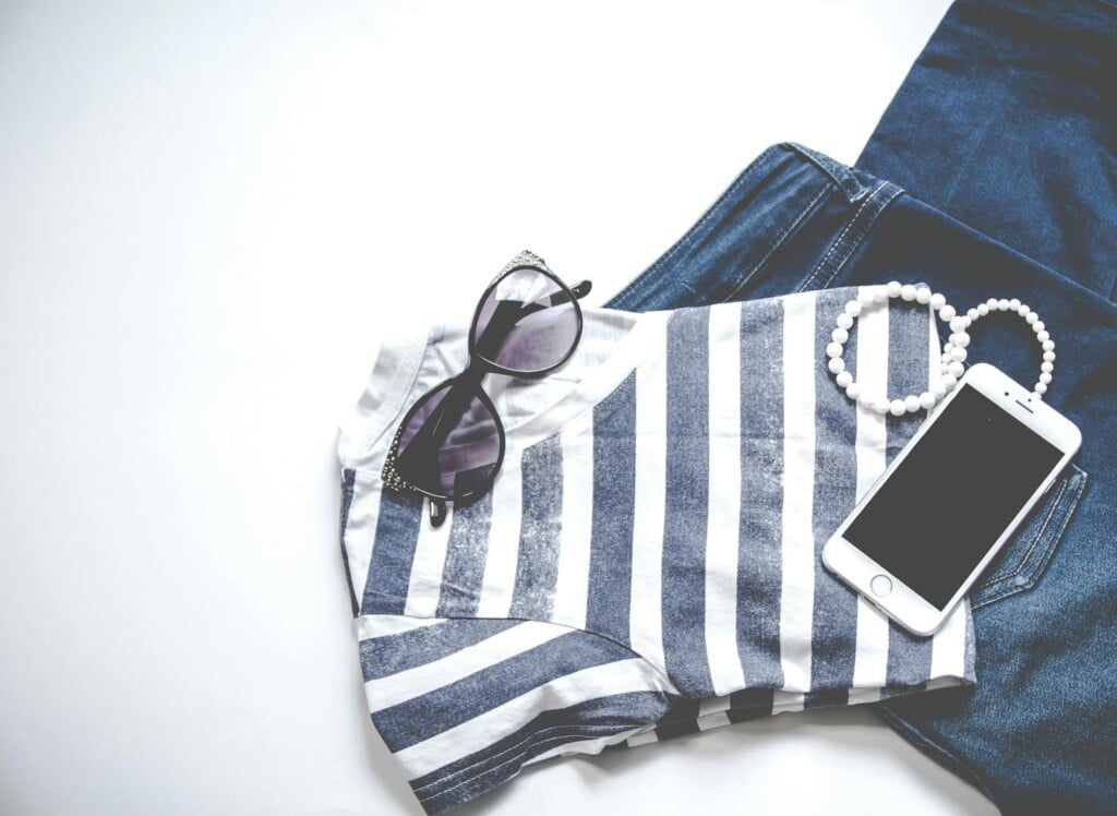 A striped shirt and blue jeans are neatly arranged on a white surface. Accessories include a pair of sunglasses, a white beaded bracelet, and a smartphone, creating a casual fashion ensemble.