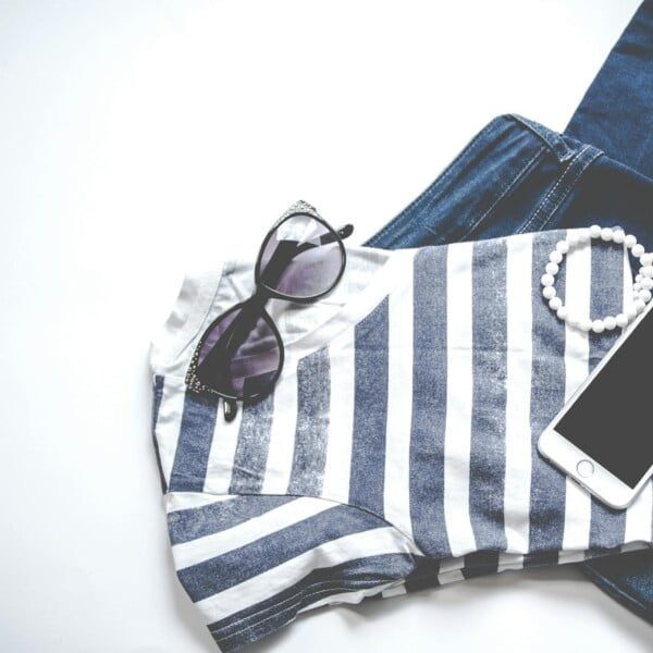 Capsule Wardrobe Essentials for Women Over 40 38 A striped shirt and blue jeans are neatly arranged on a white surface. Accessories include a pair of sunglasses, a white beaded bracelet, and a smartphone, creating a casual fashion ensemble.