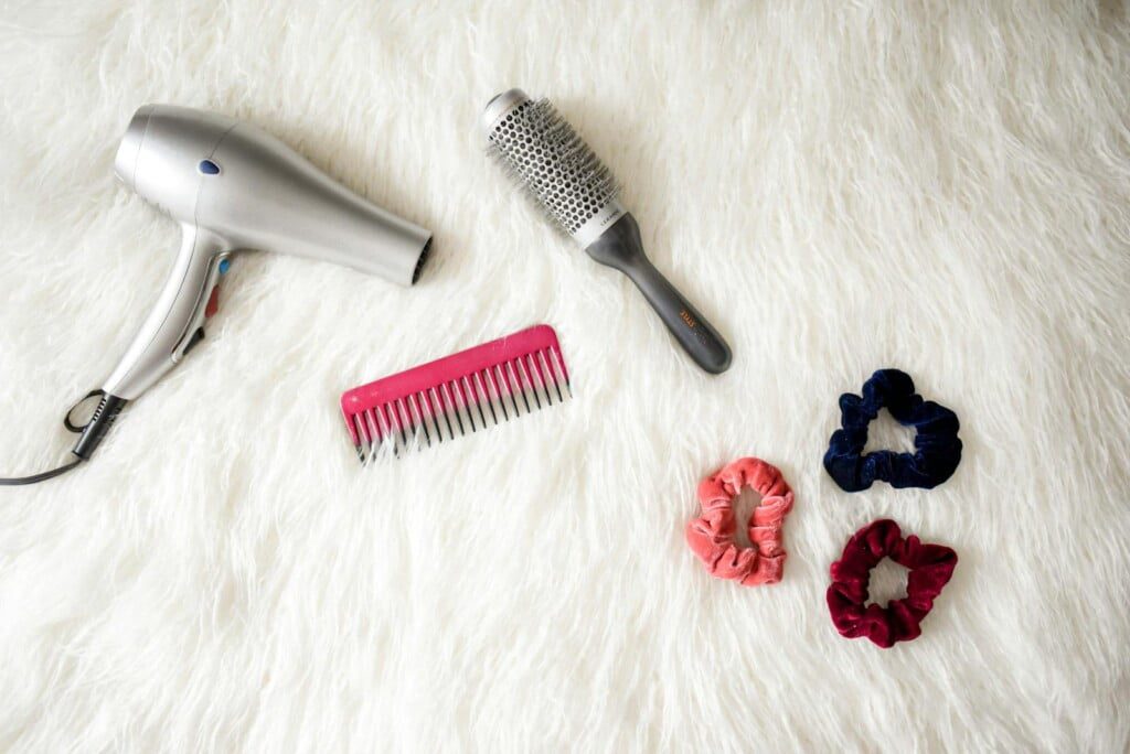 A silver hairdryer, a round brush, a pink comb, and three vibrant scrunchies in blue, pink, and red are arranged on a white fuzzy surface—perfect Hair Tool Essentials to regain hair health.