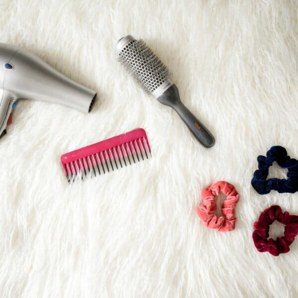 A silver hairdryer, a round brush, a pink comb, and three vibrant scrunchies in blue, pink, and red are arranged on a white fuzzy surface—perfect Hair Tool Essentials to regain hair health.