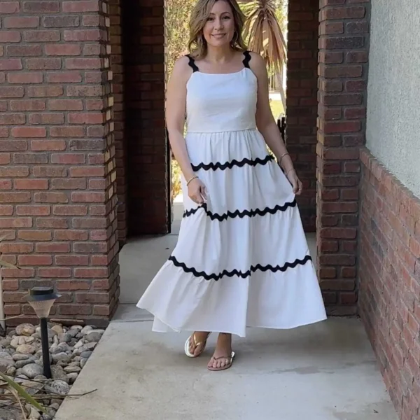 Summer Dresses for Women Over 40 for the Hot Days Ahead 15 A woman stands outdoors in a brick archway, wearing a flowing white dress with black scalloped trim and sandals. She smiles at the camera, with greenery visible in the background.