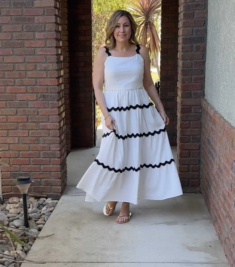 A woman stands outdoors in a brick archway, wearing a flowing white dress with black scalloped trim and sandals. She smiles at the camera, with greenery visible in the background.