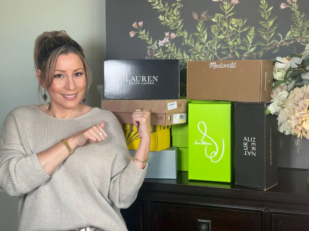 A woman smiling and pointing at a stack of branded shoe boxes on a table. The background features floral wallpaper, and there are various shoes inside the boxes. Floral decor is visible on the right.