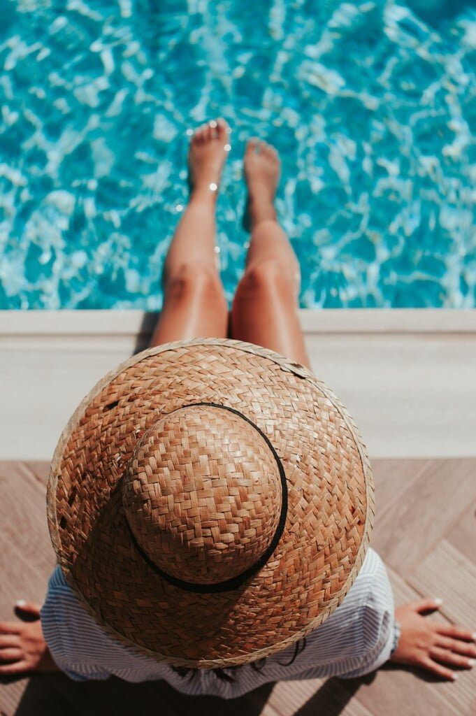 A person in a stylish summer dress and straw hat sits by the edge of a swimming pool, their legs dipped in the sparkling water. The scene captures a relaxing, sunny day, perfect for adding new finds from SHEIN to your closet.