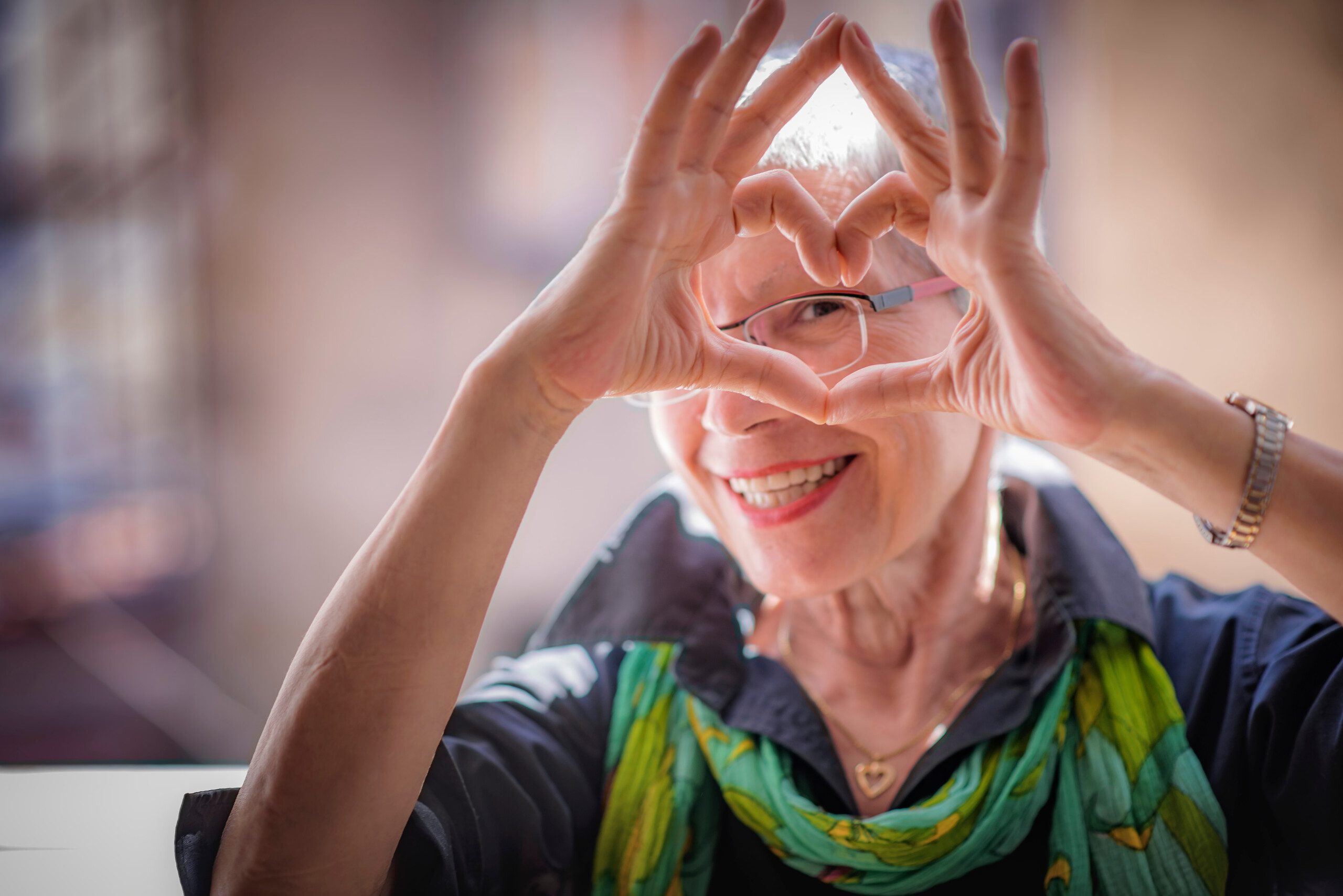 An older woman smiling and making a heart shape with her hands around her eyes. She is wearing glasses, a black shirt, and a colorful scarf. The background is softly blurred.