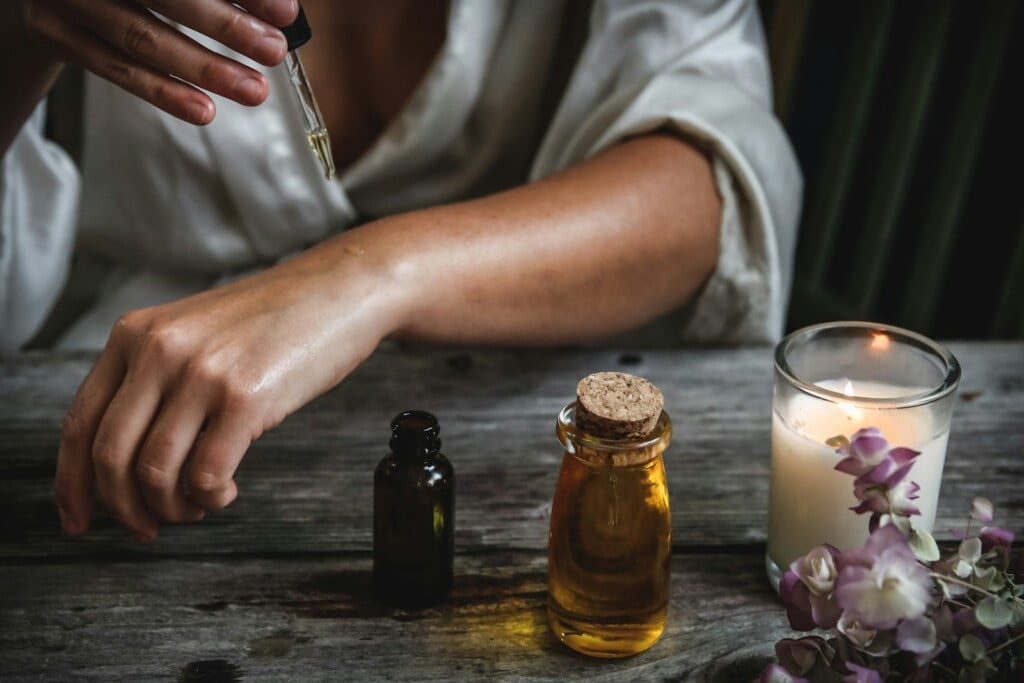 A person applies castor oil on their wrist using a dropper over a rustic wooden table. Nearby are bottles of oil, a lit candle, and purple flowers, creating a soothing ambiance.