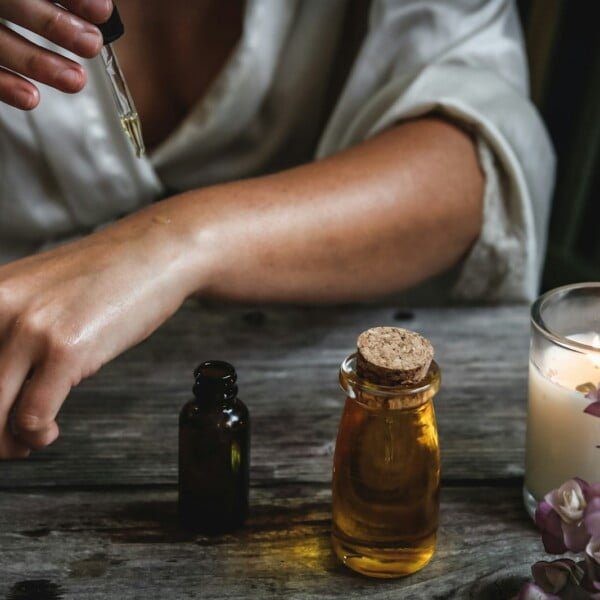 My Personal Experience With Castor Oil And Benefits I've Found 9 A person applies castor oil on their wrist using a dropper over a rustic wooden table. Nearby are bottles of oil, a lit candle, and purple flowers, creating a soothing ambiance.