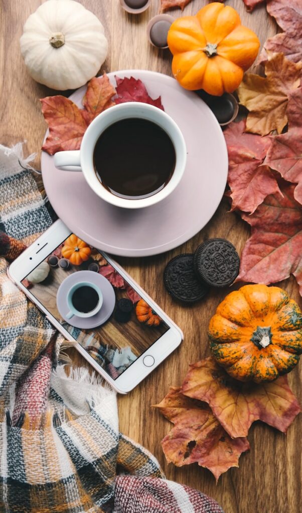 A cozy autumn scene unfolds with a cup of coffee on a pink plate, surrounded by pumpkins, colorful leaves, and Oreo cookies on a wooden table. A smartphone displays the setting, highlighting fashion over 40 and how it beautifully embraces the fall transition alongside a warm plaid scarf.