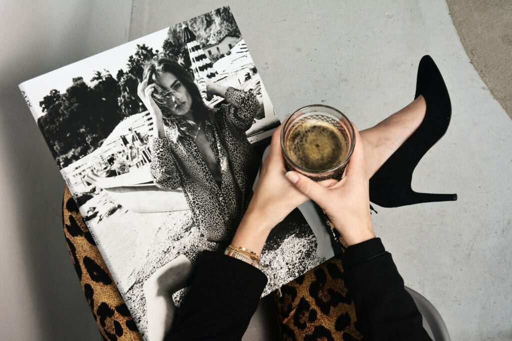 A person clad in leopard print pants and black high heels, perfectly embodying fall fashion, holds a black-and-white fashion magazine and a glass of dark liquid. The magazine showcases a woman posing outdoors, capturing the essence of the seasons style.