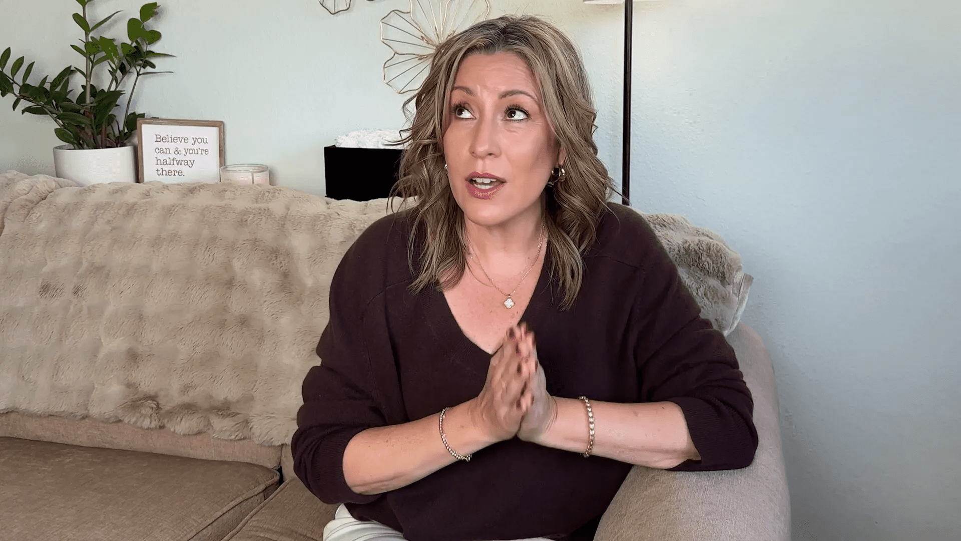 Unlocking the Secrets of Castor Oil: 5 Transformative Benefits for Aging Gracefully 37 A woman with shoulder-length hair and a dark sweater sits on a beige couch with her hands clasped, looking upward. Behind her is a decorative wall piece, a lamp, and a small sign.