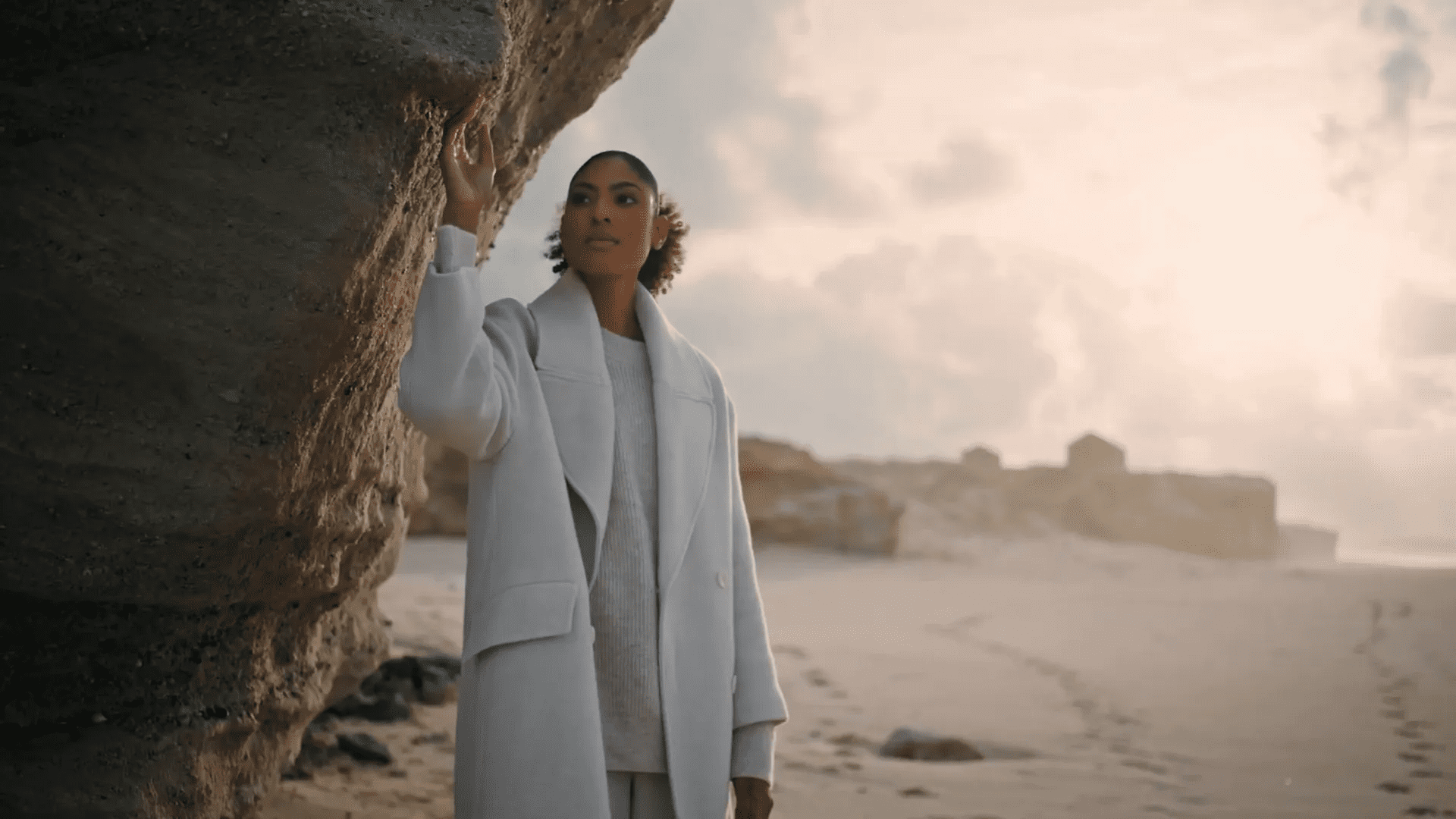 A person in a light gray coat and outfit, exemplifying how to identify quality clothing, stands by a large rock formation on a beach. They look upwards, hand touching the rock. The sky is cloudy, with footprints trailing in the sand.