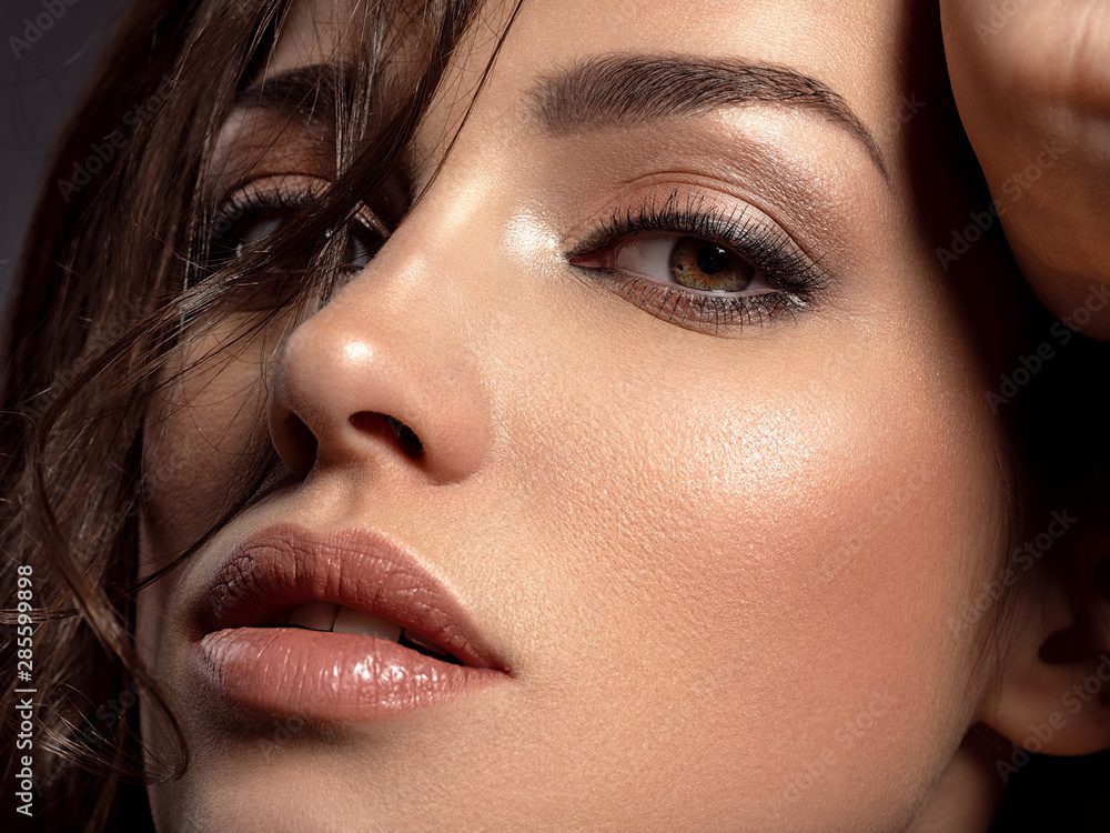 Close-up of a womans face with brown eyes and glossy lips. Her skin is smooth with subtle makeup, including eyeliner and shimmering eyeshadow in a Mocha Mousse shade. Strands of hair partially cover her forehead, creating a dramatic, intimate look inspired by 2025 fashion trends.