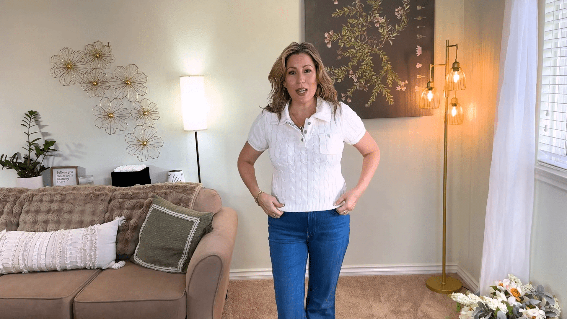 A person in a white short-sleeved shirt and blue jeans, perfect for browsing trendy stores for women over 40, stands in a living room. Behind them are a sofa, decorative floral wall art, a floor lamp with warm lights, and a window with blinds. A cushioned chair and small plant are also visible.