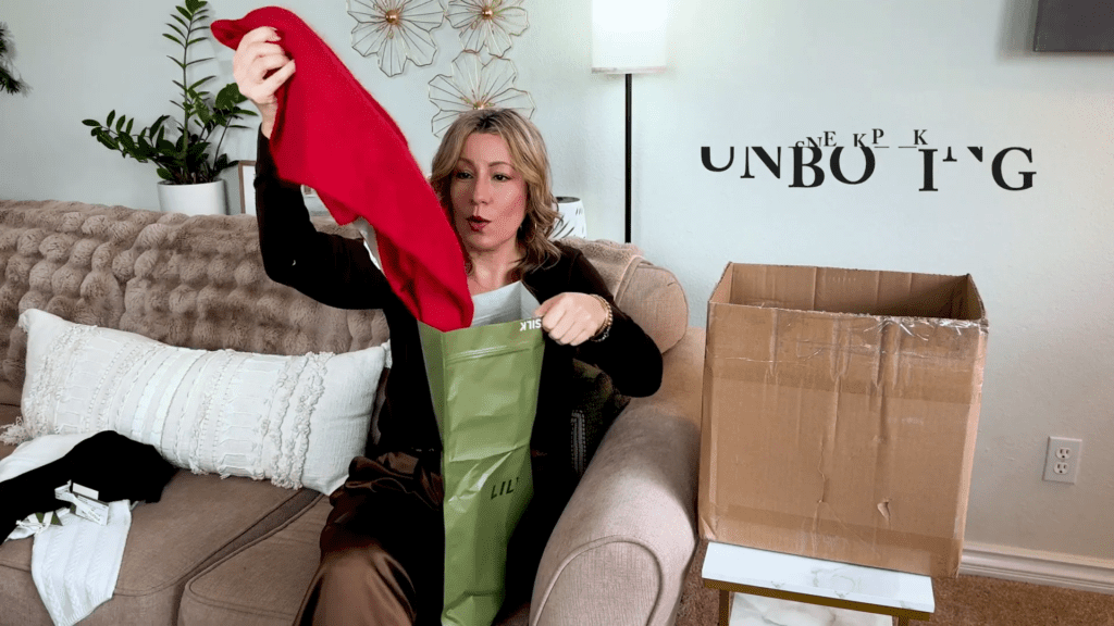 A person sitting on a couch is unboxing a red garment from a green bag. Theres an open cardboard box beside them. Wall decor and the word unboxing are visible in the background.